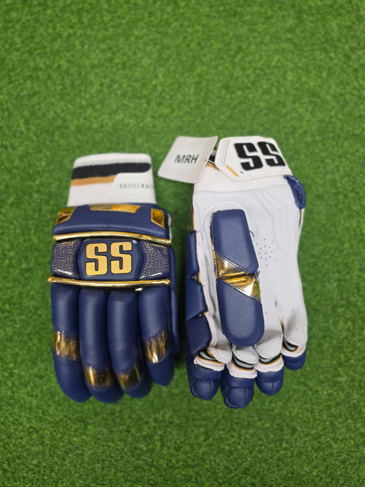 A pair of SS Super Test Mumbai Indians IPL Edition Cricket Batting Gloves in navy blue, white, and gold, featuring premium finger protection and ergonomic grip on a green grass background.