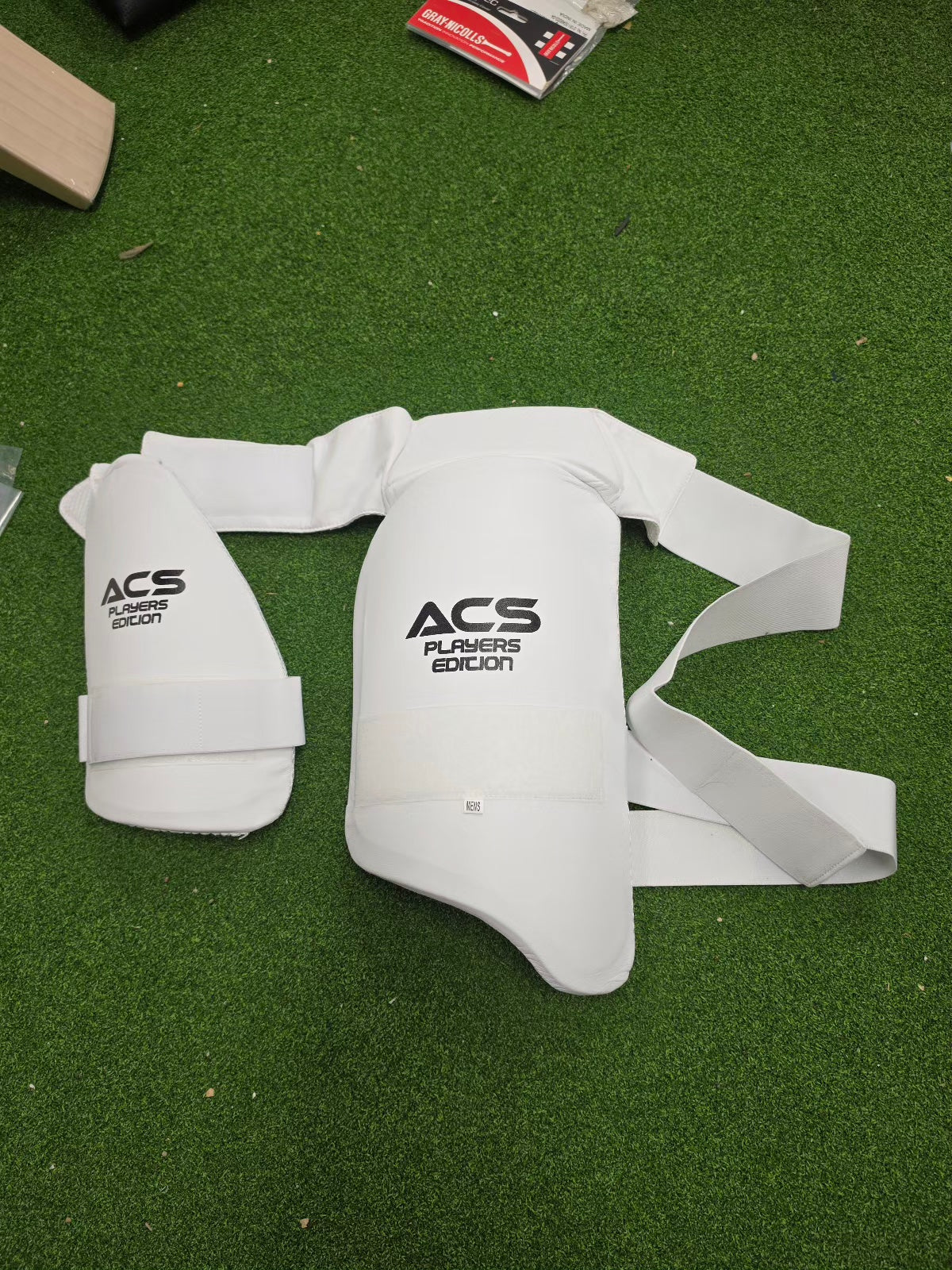 Flat lay view of white ACS Superlite Cricket Double Thigh Pads for left hand batsmen on a green turf surface from Albertacricketstore.com.