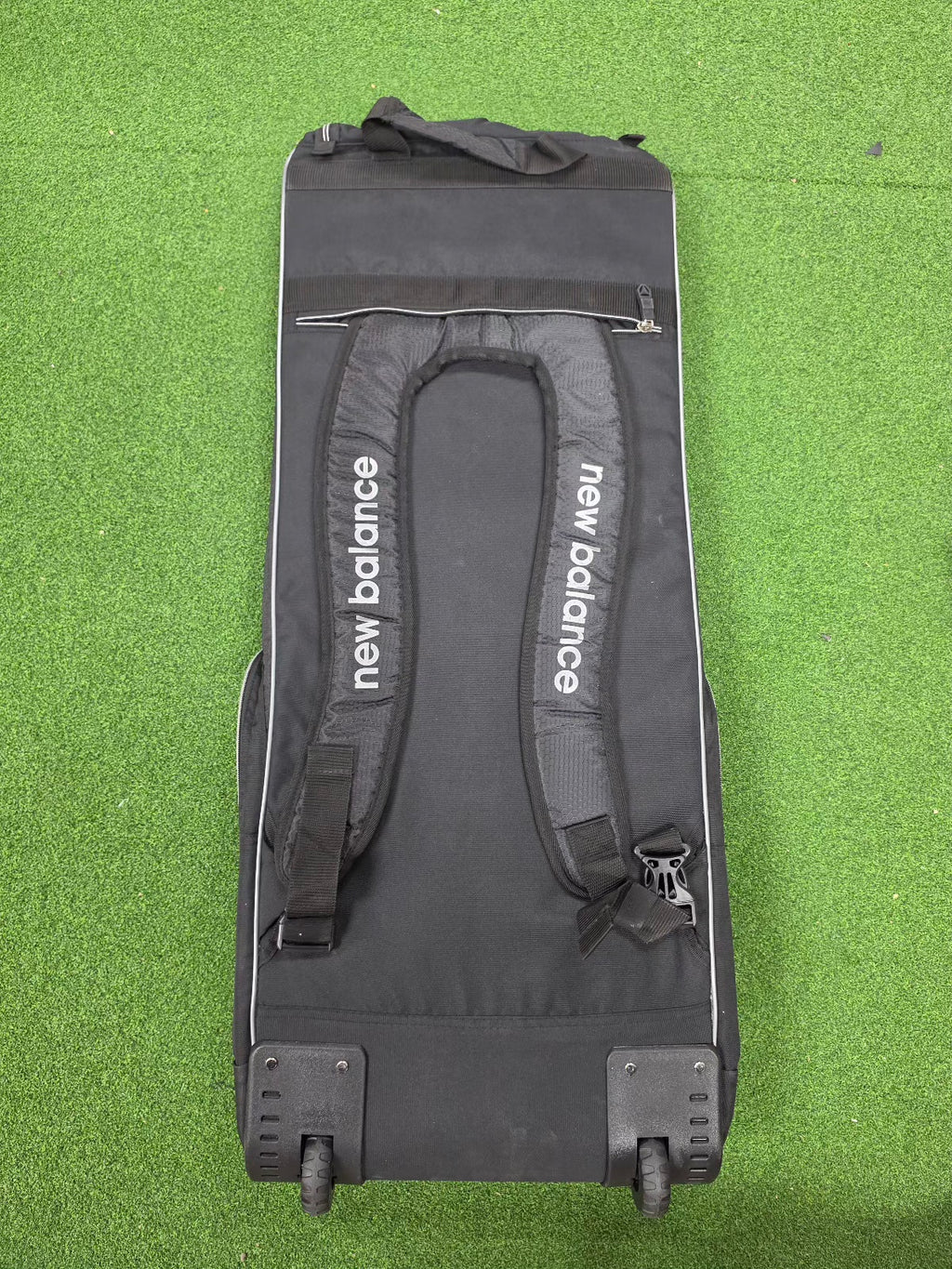 Overhead view of a black New Balance 10i combo duffle wheelie cricket bag with heavy-duty tractor wheels and backpack straps on green grass.