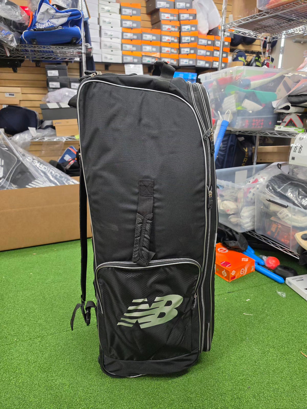 Side view of a black New Balance 10i combo duffle wheelie cricket bag with silver branding standing in a sports store.