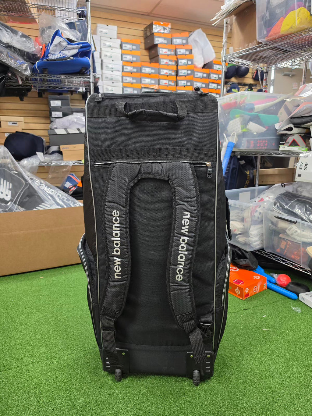 Rear view of black New Balance 10i combo duffle wheelie cricket kit bag showing padded backpack shoulder straps standing in a shop.