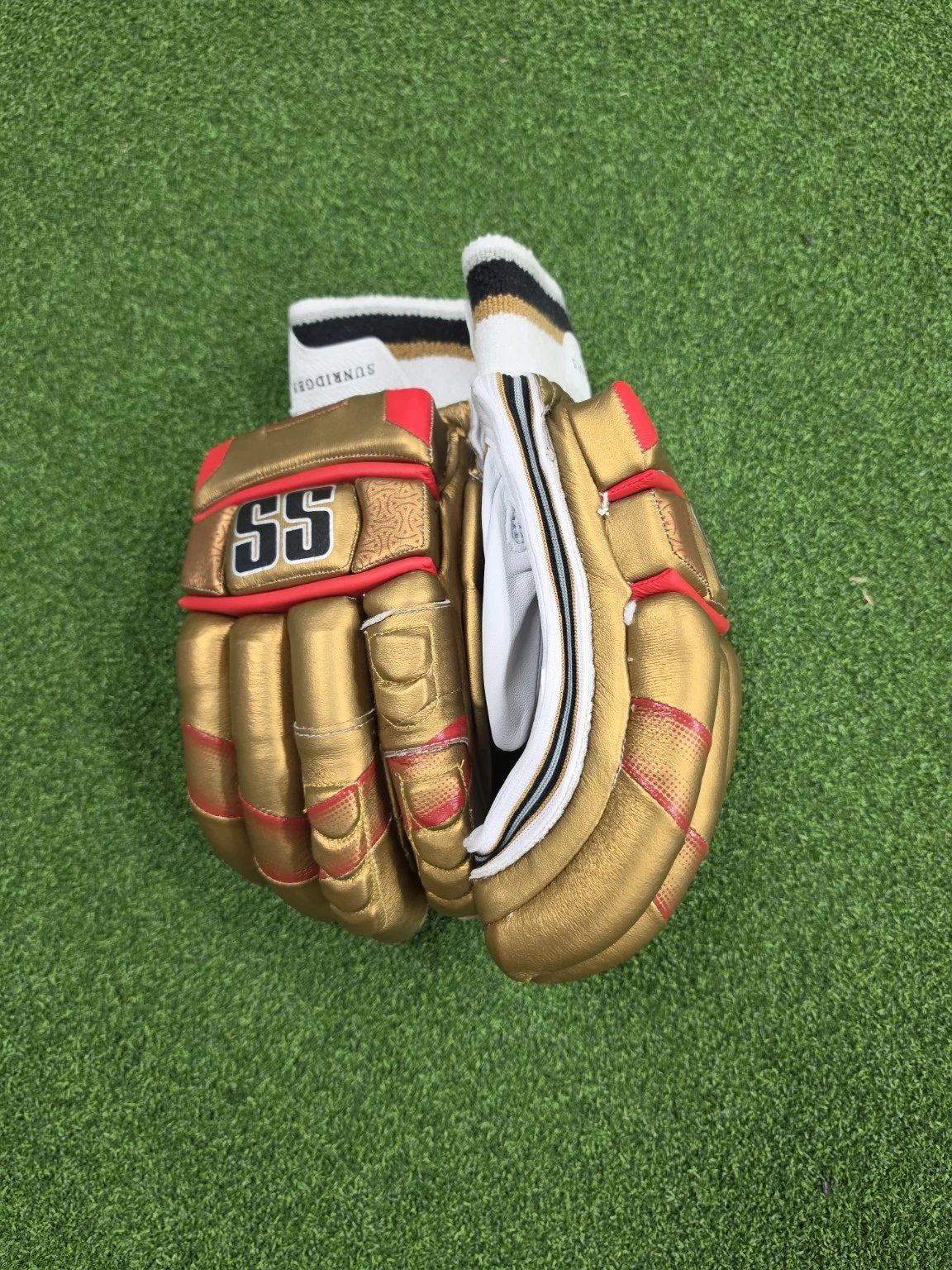 Close up top view of gold, red, and white SS Super Test Royal Challengers Bangalore IPL cricket batting gloves on artificial turf