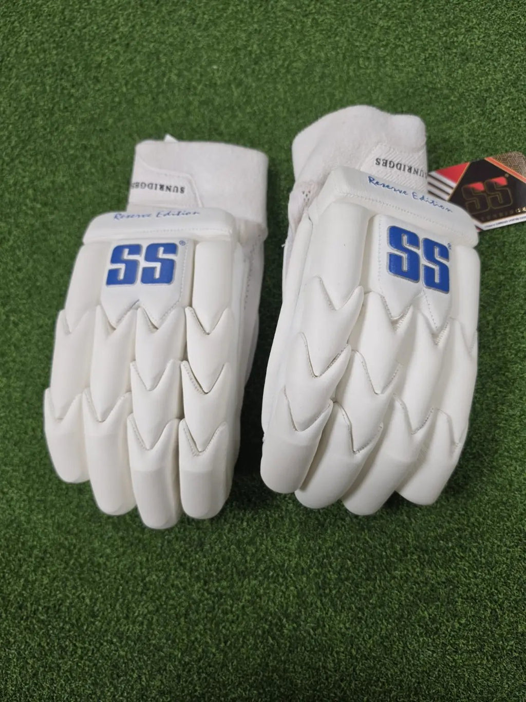 Pair of white SS Ton Reserve Edition cricket batting gloves with blue SS logos placed on artificial turf