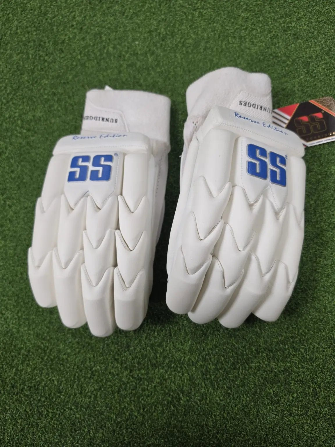 Pair of white SS Ton Reserve Edition cricket batting gloves with blue SS logos placed on artificial turf