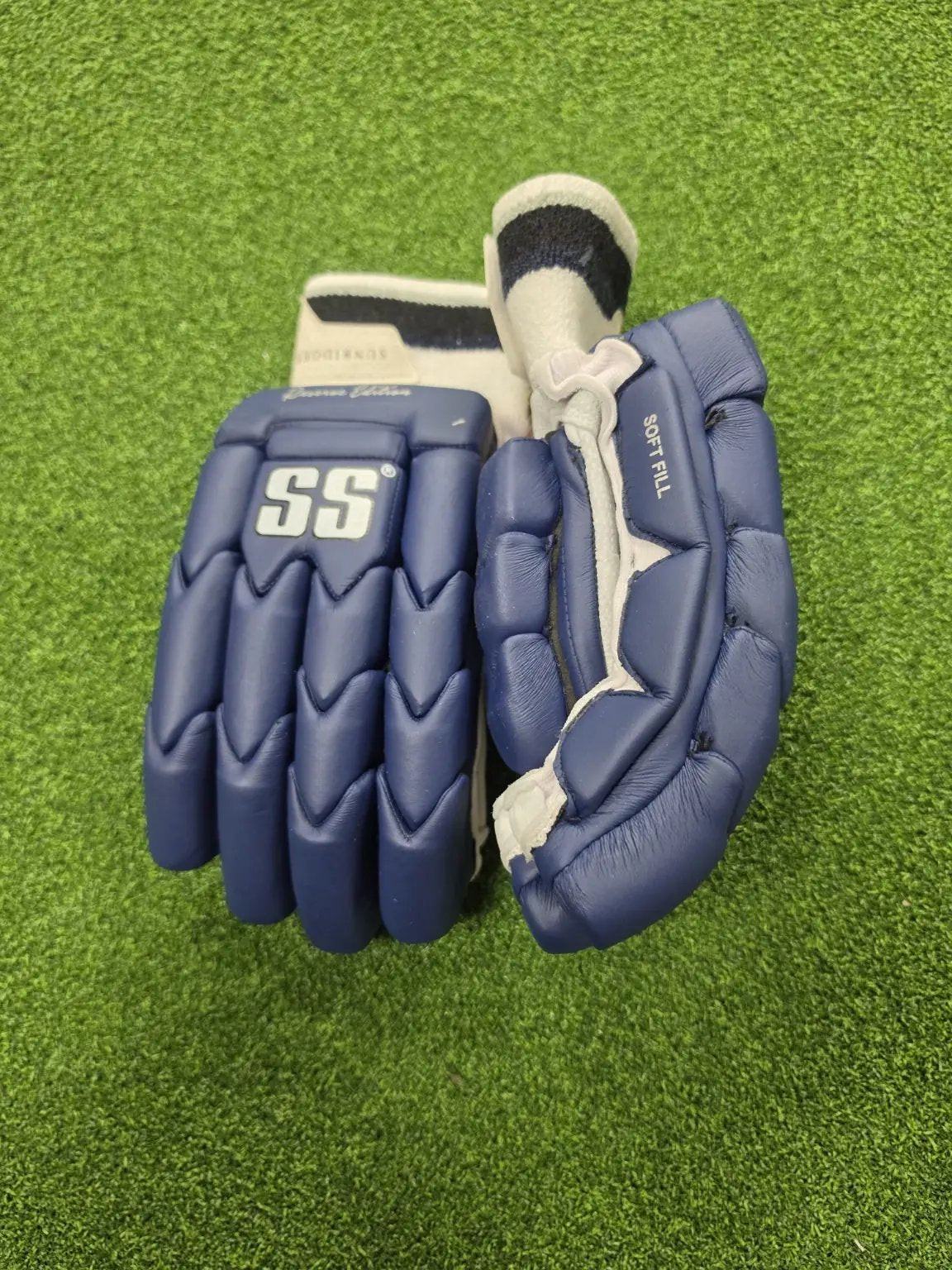 Pair of navy blue SS Ton Reserve Edition cricket batting gloves showcasing side finger protection and soft-fill thumb padding on green turf