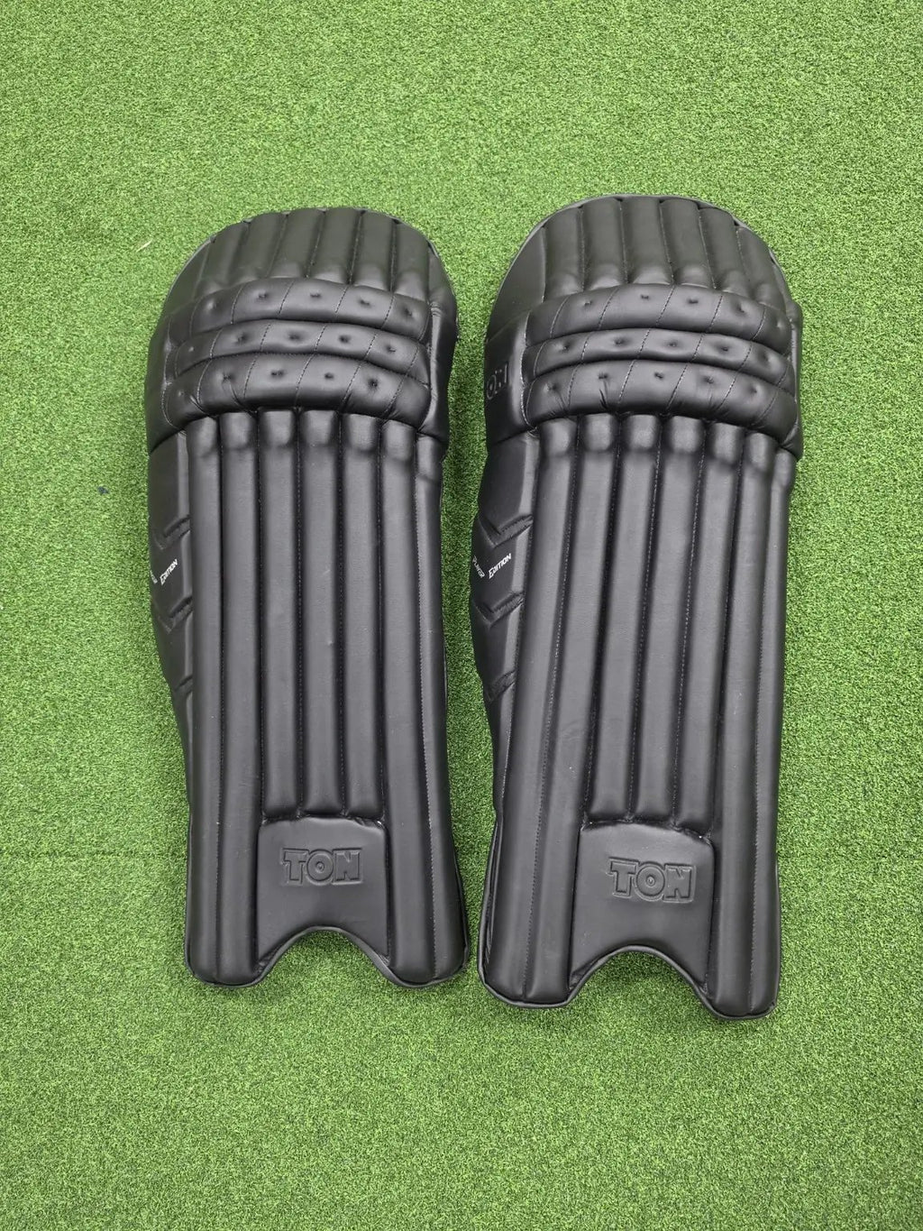Pair of TON Player Edition Black Cricket Batting Pads displayed on green grass against a transparent lower border.