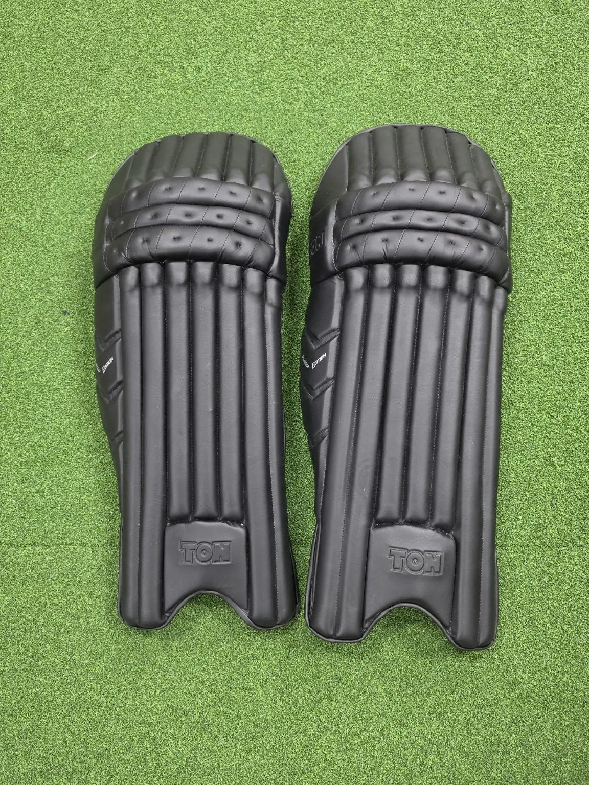 Pair of TON Player Edition Black Cricket Batting Pads displayed on green grass against a transparent lower border.