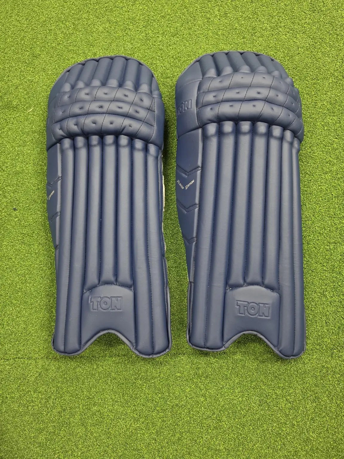 Top portion view of TON Player Edition Navy Blue Cricket Batting Pads showing the top knee rolls over green turf and a transparent background.