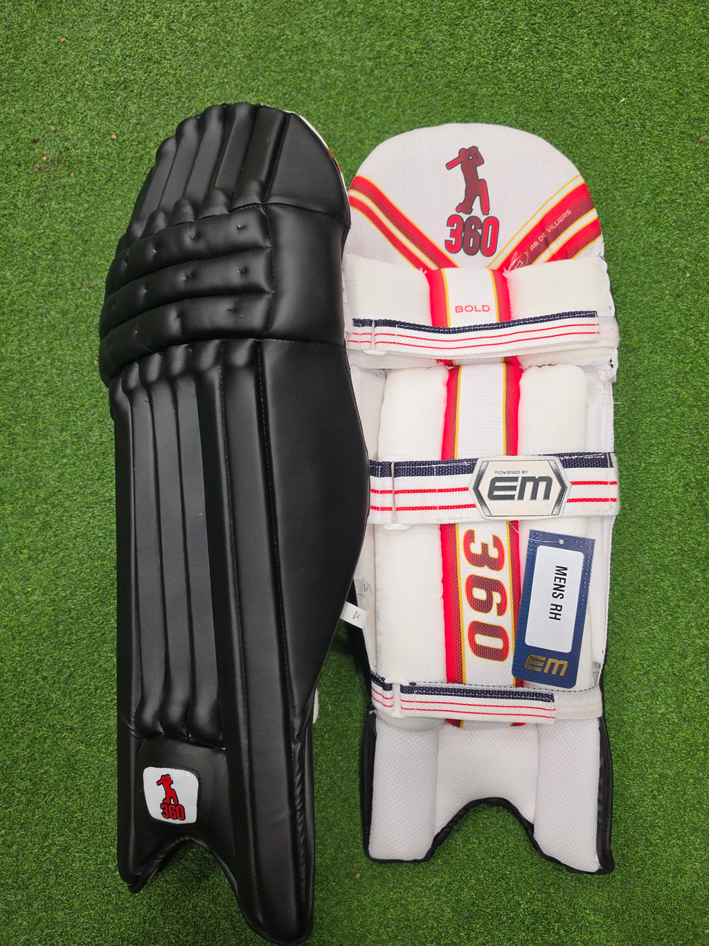 Front and inner back view of EM 360 Bold black cricket batting pads with red accents on green artificial turf.