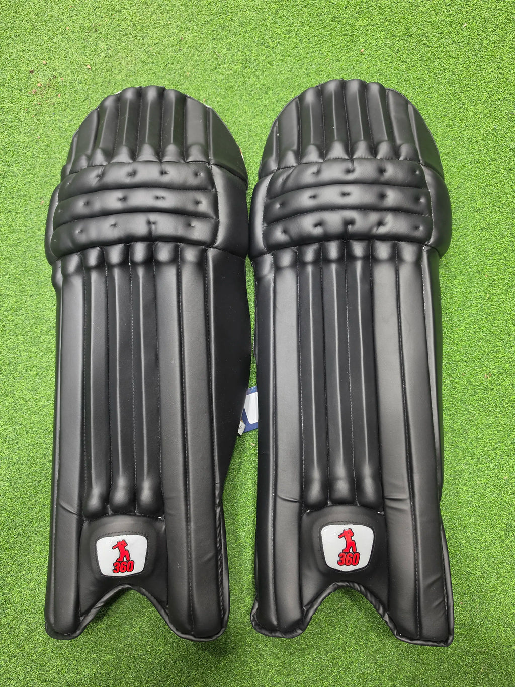 Front view of a pair of EM 360 Bold black cricket batting pads on green artificial turf.