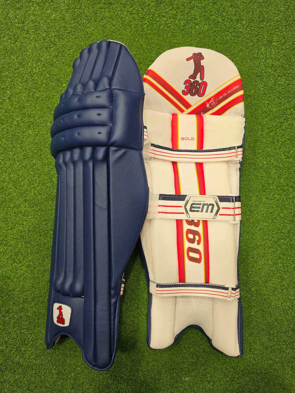 Front face and inner padded back view of EM 360 Bold adult navy blue cricket batting pads with red accents on artificial grass on Alberta.