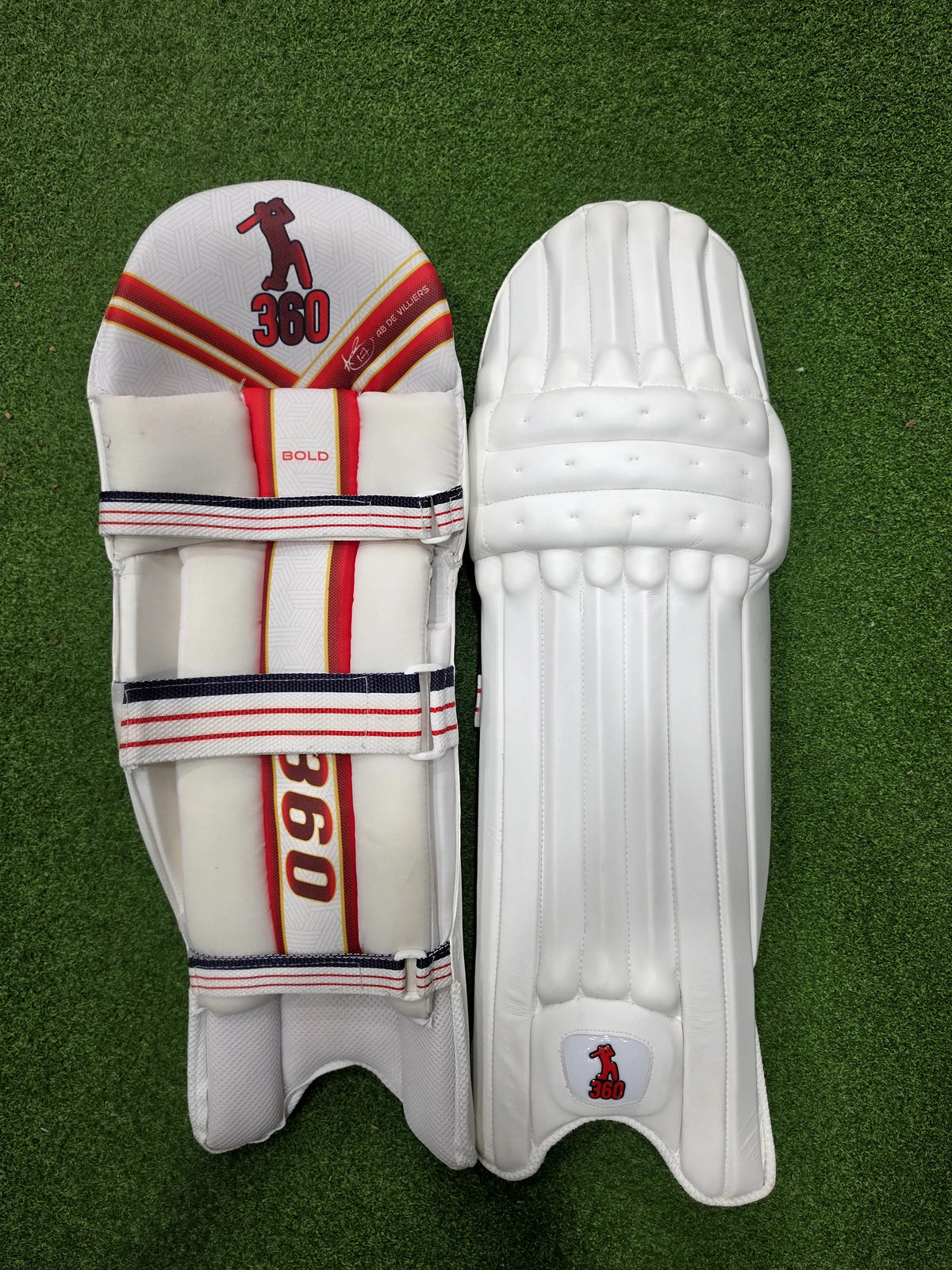 Front and inner back view of EM 360 Bold white cricket batting pads with red accents on green artificial turf.