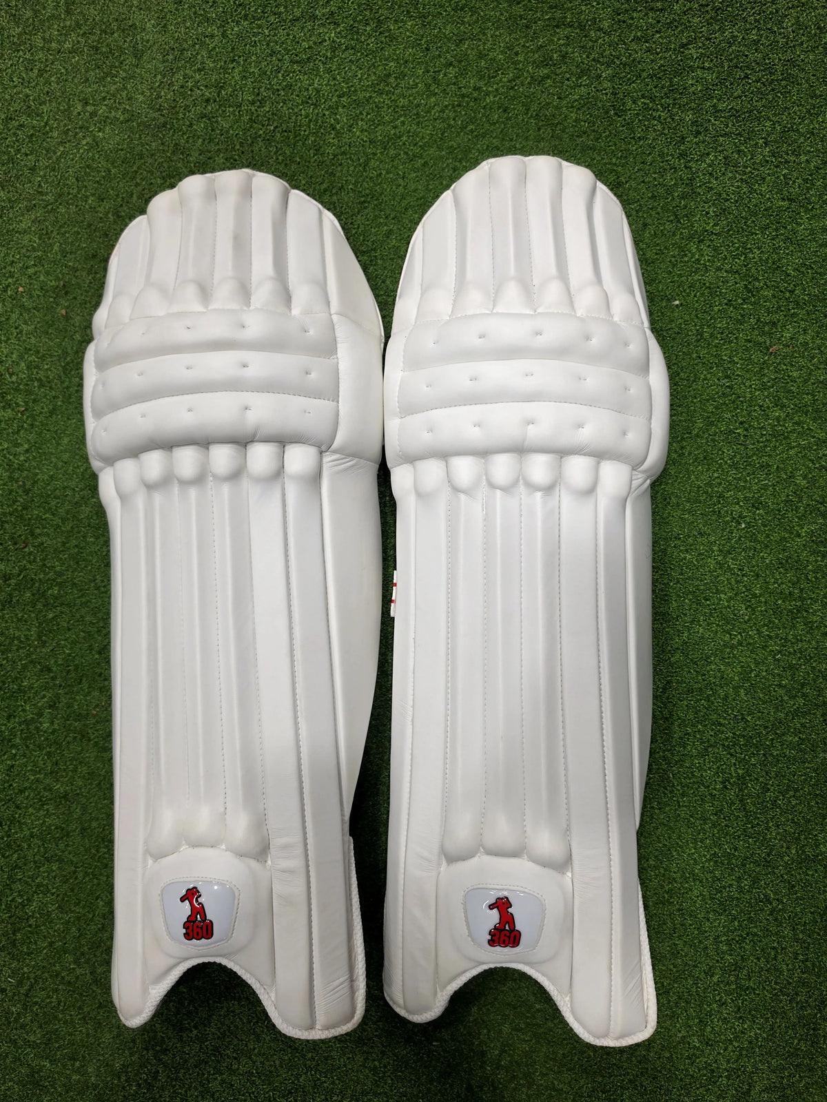 Front view of a pair of EM 360 Bold white cricket batting pads on green artificial turf.