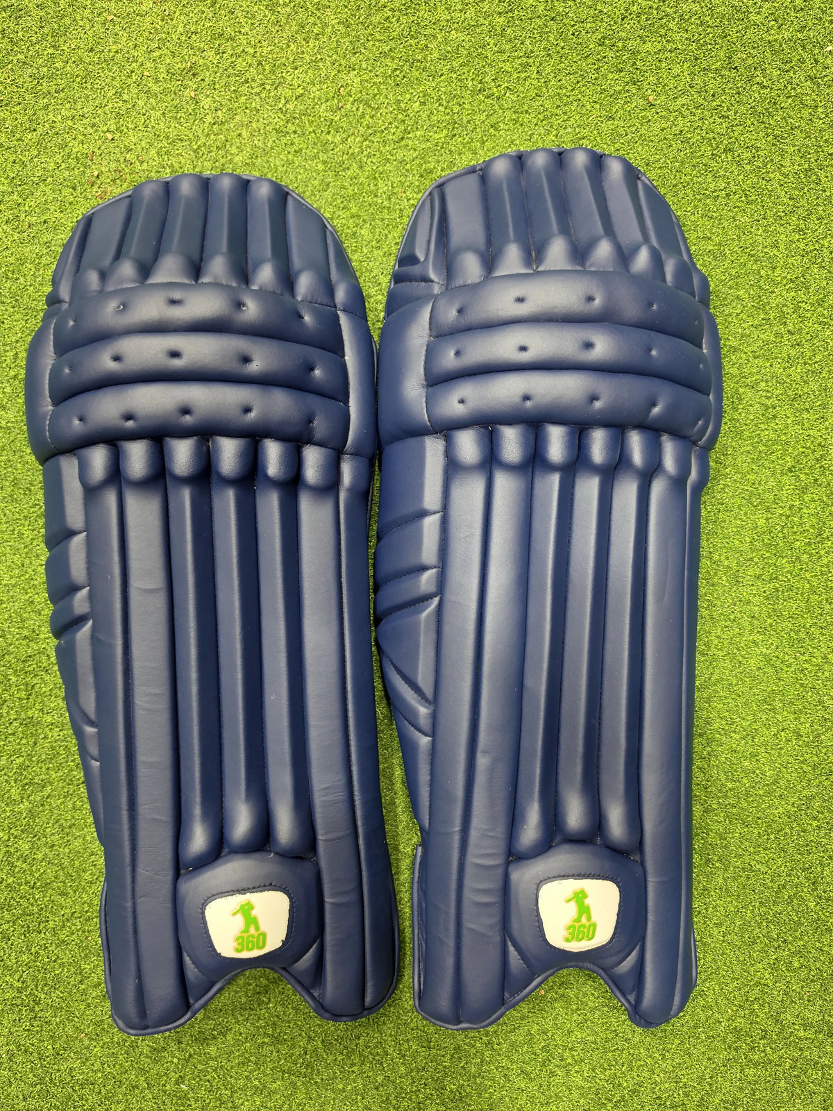 Top view of EM 360 Bull Ring adult navy blue cricket batting pads on artificial grass on Alberta.