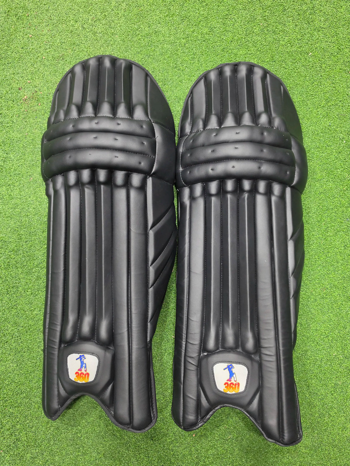 Front view of a pair of EM 360 Play Burst black cricket batting pads on green artificial turf.