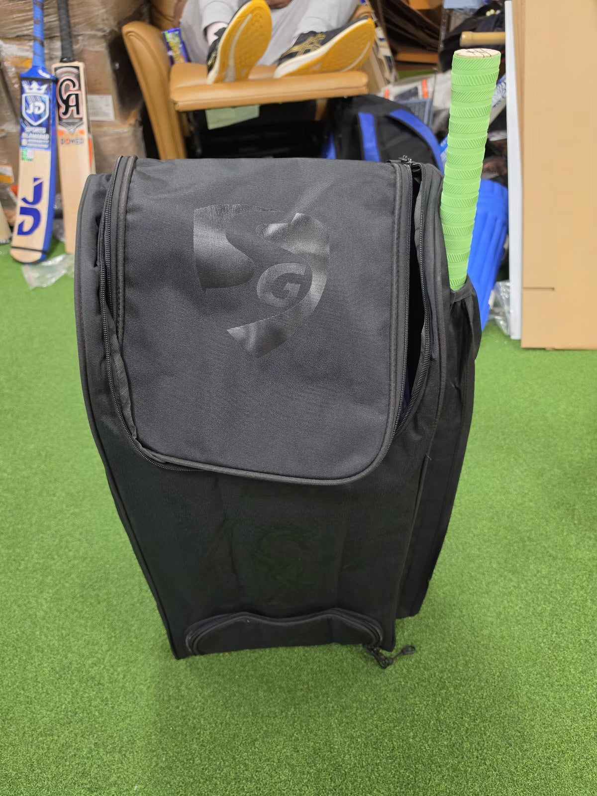 Black SG cricket kit bag standing on green turf with a green-grip bat in its side pocket.