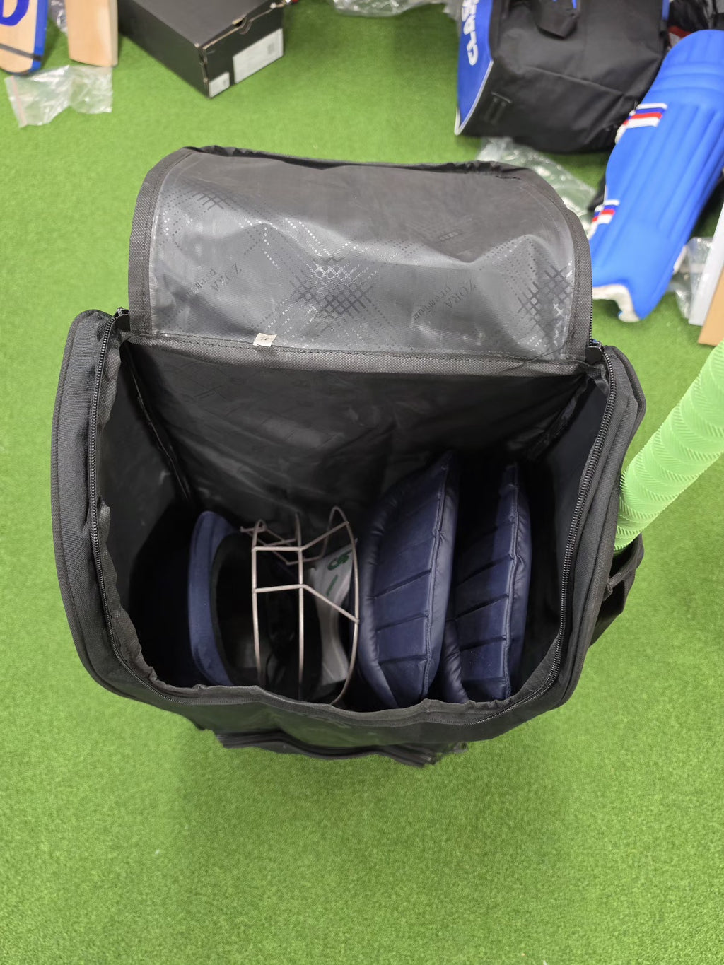 Open black cricket kit bag with navy batting pads and a helmet inside, viewed from above.