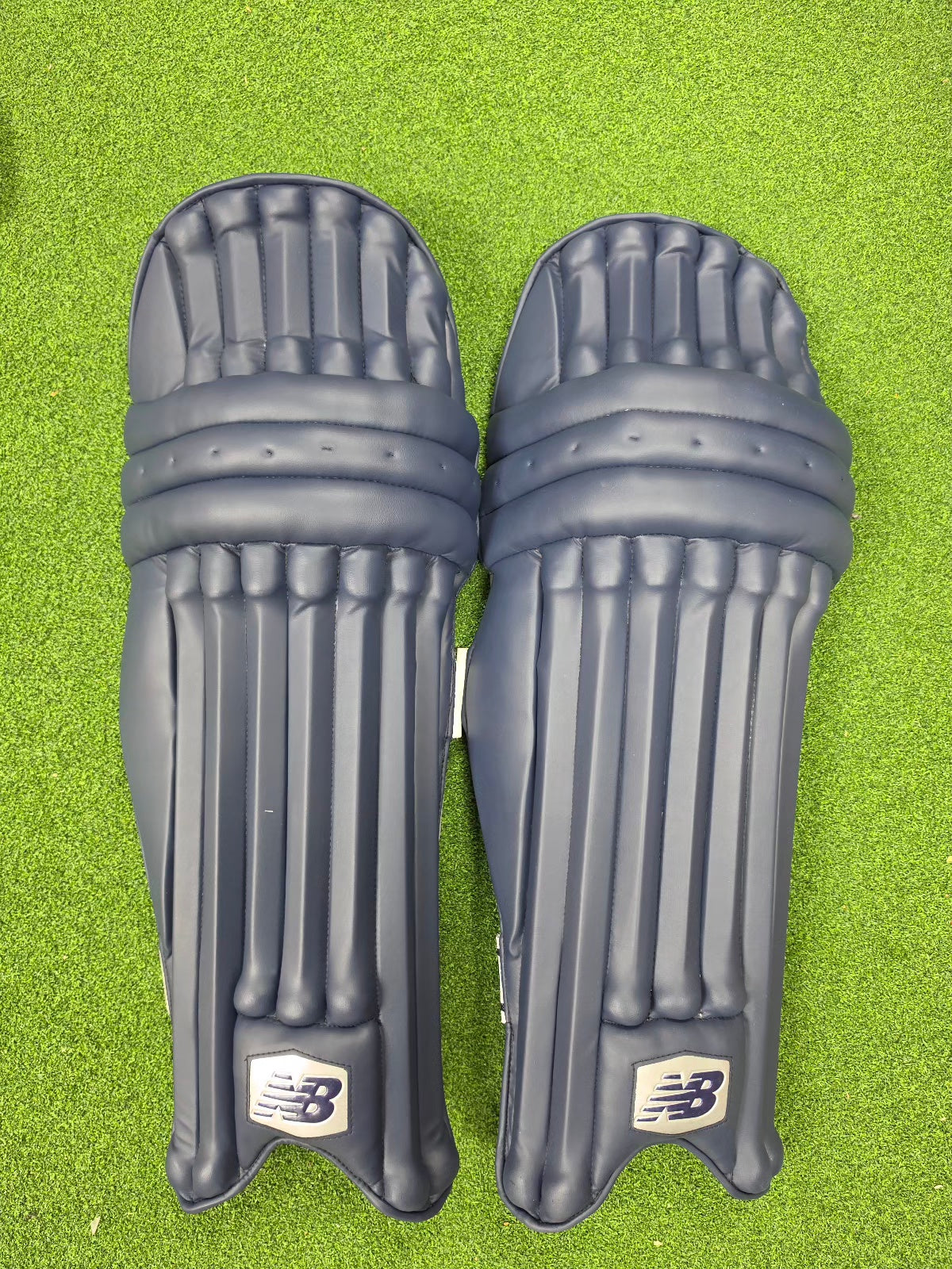 Front view of a pair of navy blue New Balance TC 8i batting pads on green turf.
